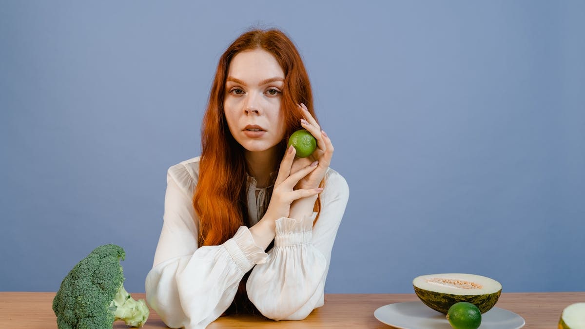 Woman with red hair holding lime surrounded by fresh produce on table.